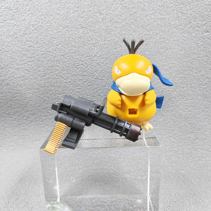 Yellow duck-like figure with a blue scarf standing beside a gray miniature Gatling gun on a clear pedestal.