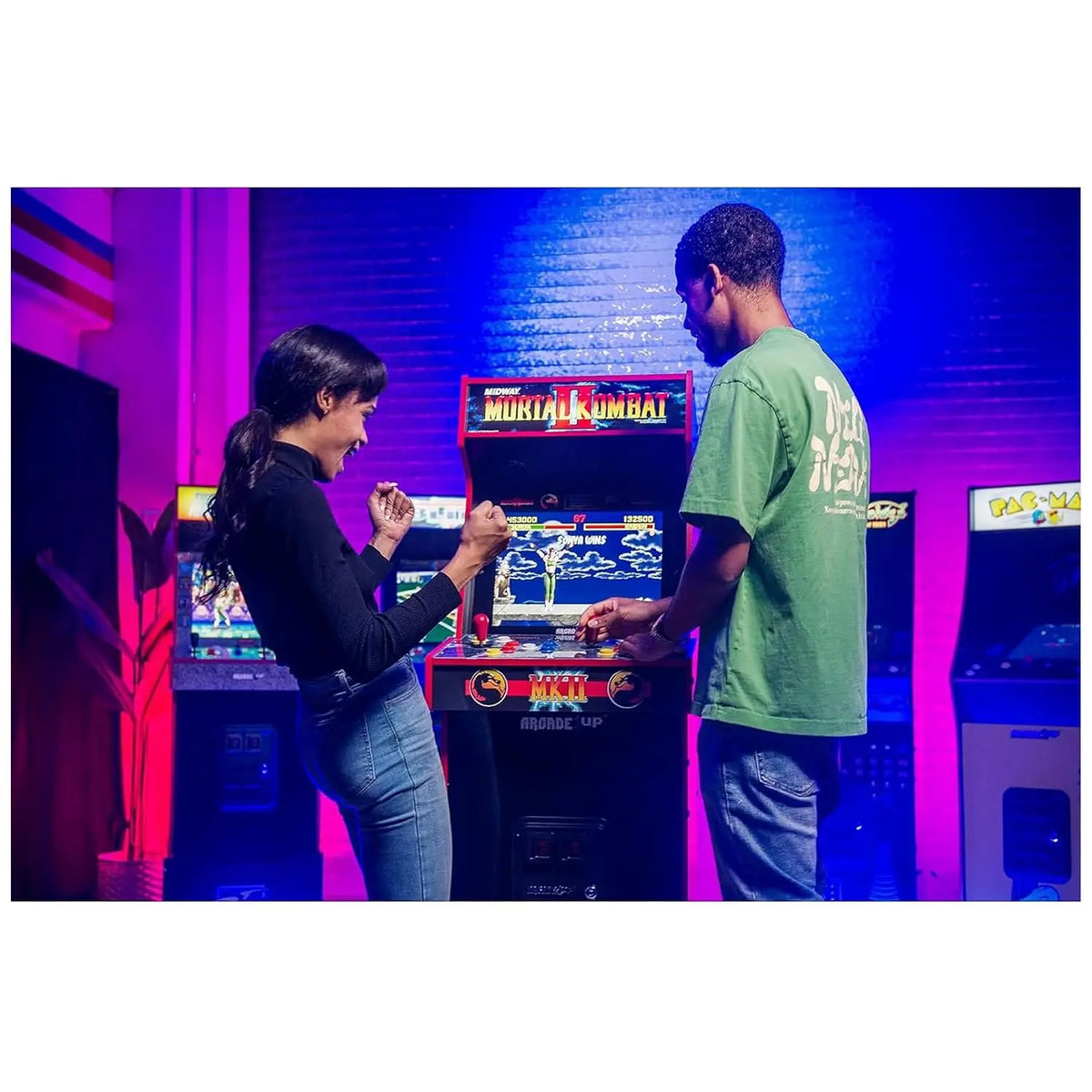 A man and woman play a Mortal Kombat II arcade machine, glowing in purple and blue light.