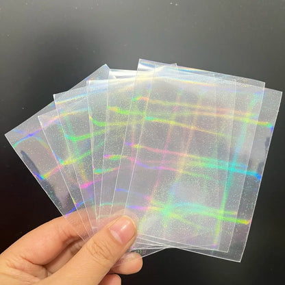 A hand holds a fan of clear trading card sleeves with a holographic laser grid and sparkle pattern.