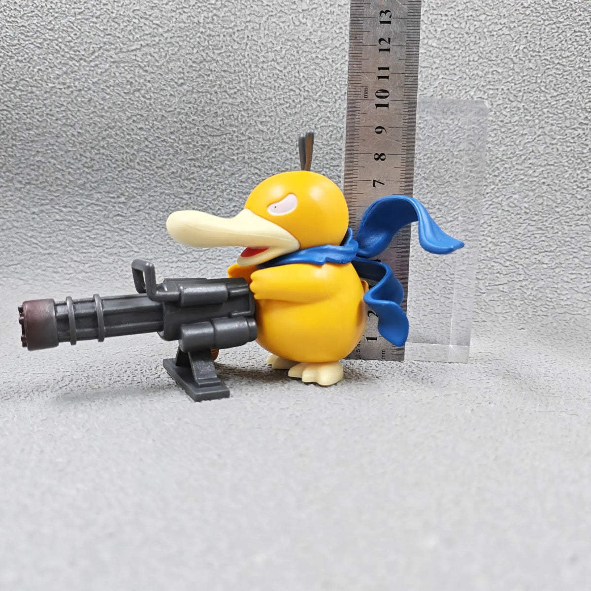 Yellow duck figurine with blue scarf, holding a large grey Gatling gun.